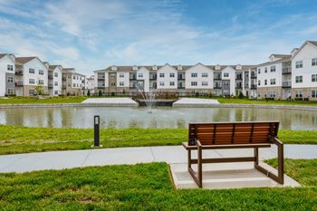 A bench sits in front of a pond with apartment buildings in the background.